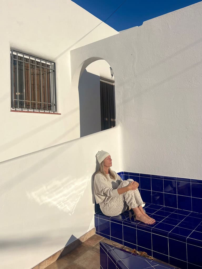 Woman in a light gray robe and white beanie sits barefoot on a blue-tiled pool edge, leaning against white walls with bright sunlight and an arched doorway in the background.
