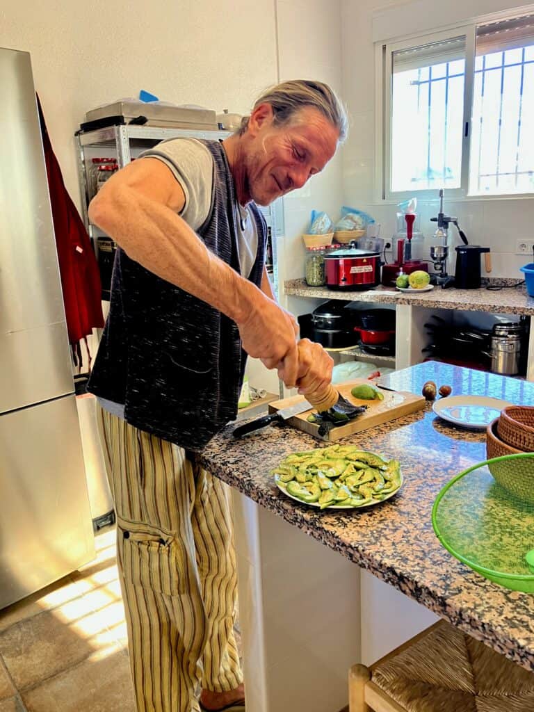 Man in a kitchen slices avocado on a wooden cutting board, plate of avocado slices nearby, wearing striped pants and a vest.