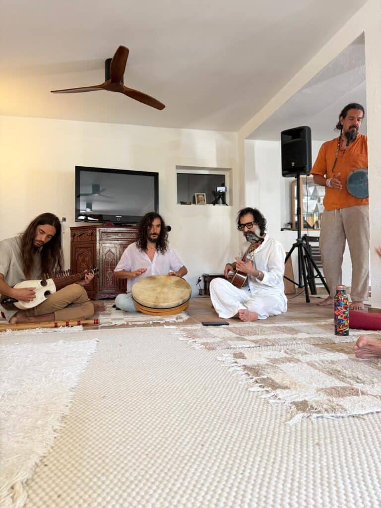 Four musicians sit on a beige rug in a bright living room, playing stringed instruments and a drum together.