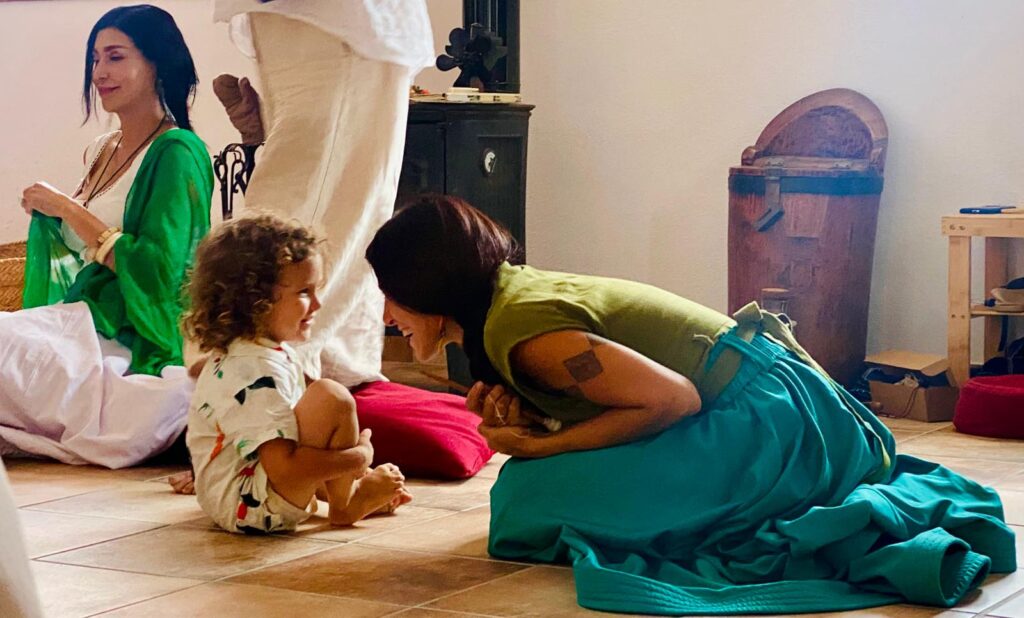 Woman in green top kneels on the floor, smiling at a curly-haired child who sits facing her, indoors with others in the background.