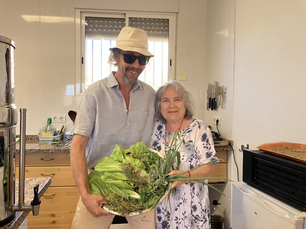 Smiling man in a beige hat and sunglasses stands beside a woman in a floral dress, holding a large leafy bouquet in a kitchen.