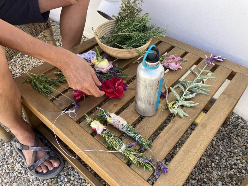 Person in sandals creating a flower garland on a wooden outdoor table with stems and greenery nearby.