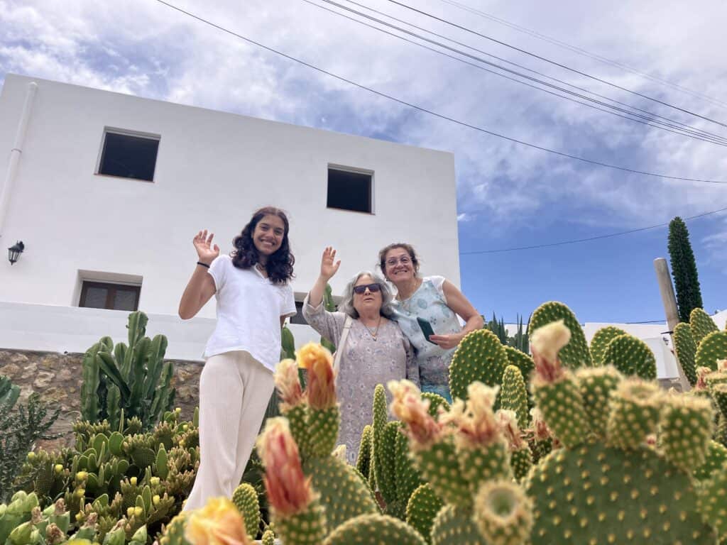 Group of smiling people waving from a garden with tall cacti in foreground and a white modern house against a blue sky.