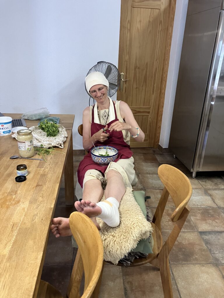 Smiling woman wearing a head scarf and red apron sits at a kitchen table, cutting herbs while a boot casts her leg in a wrap.