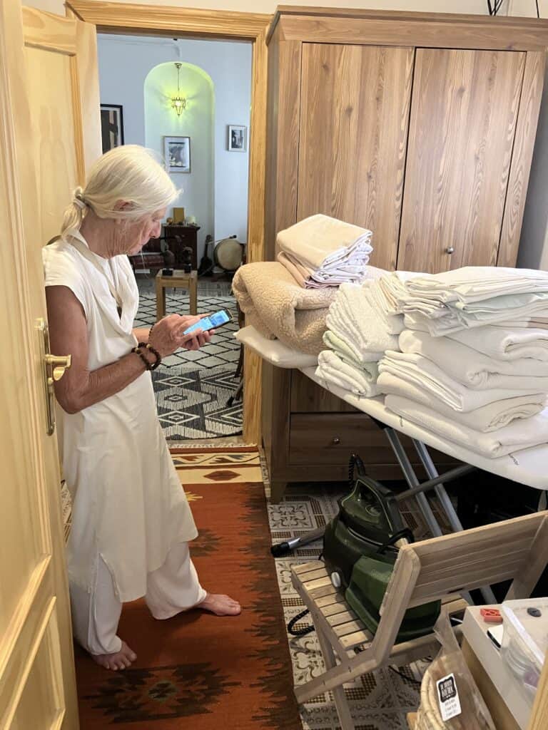 Elderly woman with white hair in a white outfit, barefoot, looking at a smartphone beside a stack of folded white linens on an ironing board in a closet/laundry area, with a wooden wardrobe nearby.