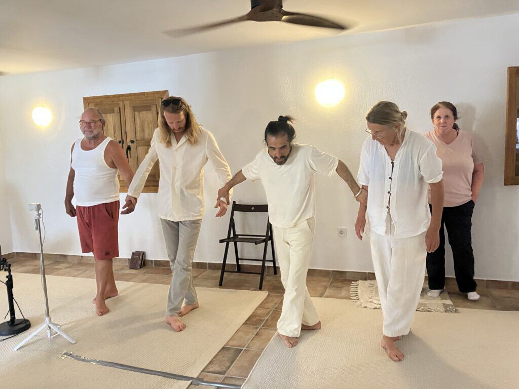 Group of adults in a living room doing a balance exercise, holding hands while standing on mats; barefoot, wearing white outfits.
