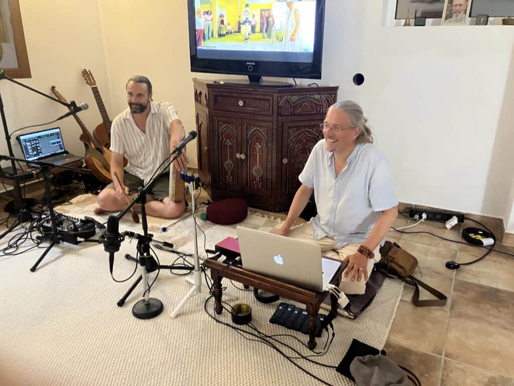 Two men recording a podcast in a home studio, with microphones, laptops, and audio gear, a guitar in the corner and a TV on an ornate cabinet behind them.