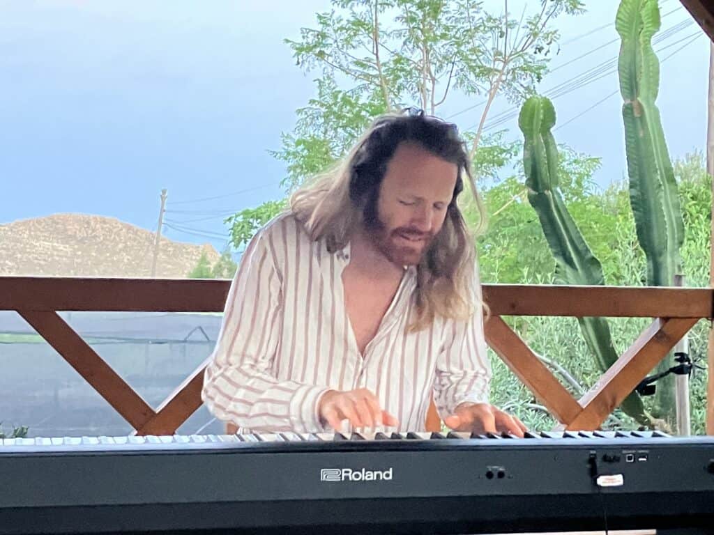 Man with long hair wearing a striped shirt plays a Roland keyboard outdoors in sunlight.