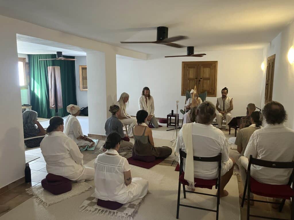 Group of people in white robes seated on cushions in a bright room, practicing meditation or ceremony.