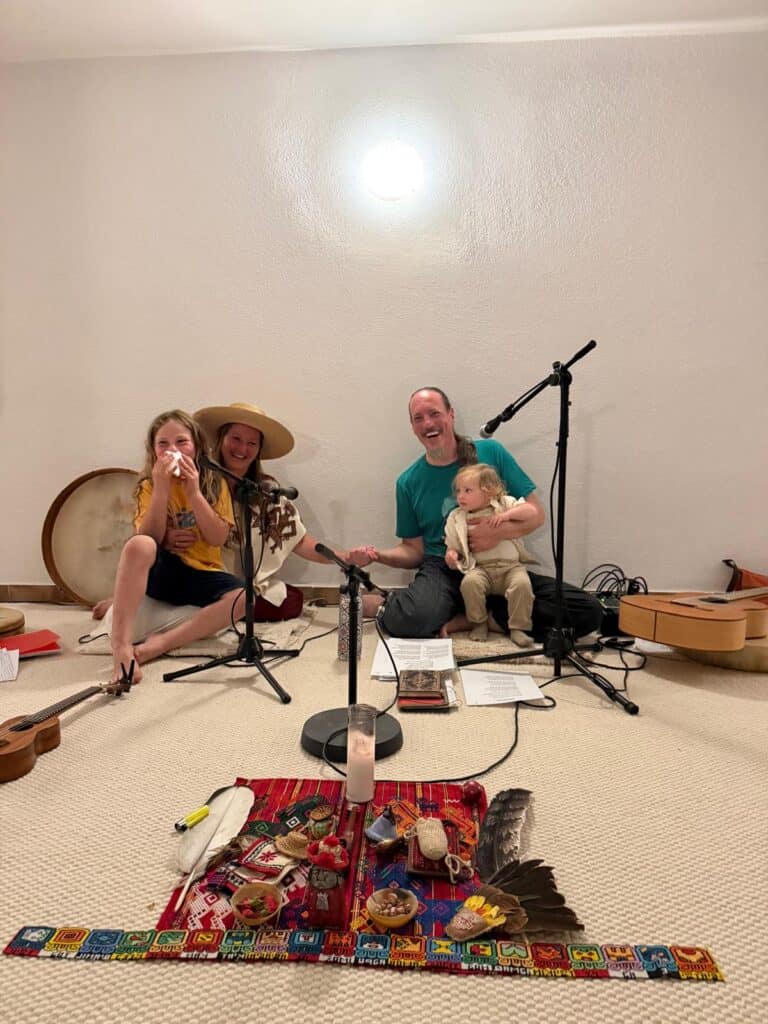 A family music session: a man holding a young child sits with two women on a beige carpet, with microphones and a guitar nearby, smiling at the camera in a simple room.